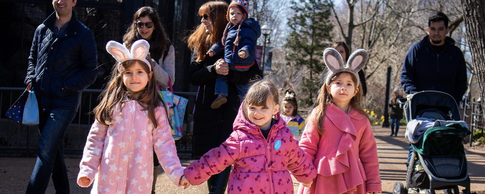 Young girls holding hands wearing bunny ear hats with parents in the background.