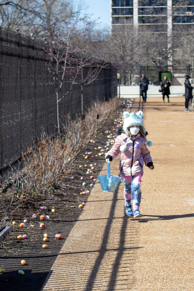 Young girl with winter hat and mask carrying an Easter basket with colored, plastic eggs on the ground.