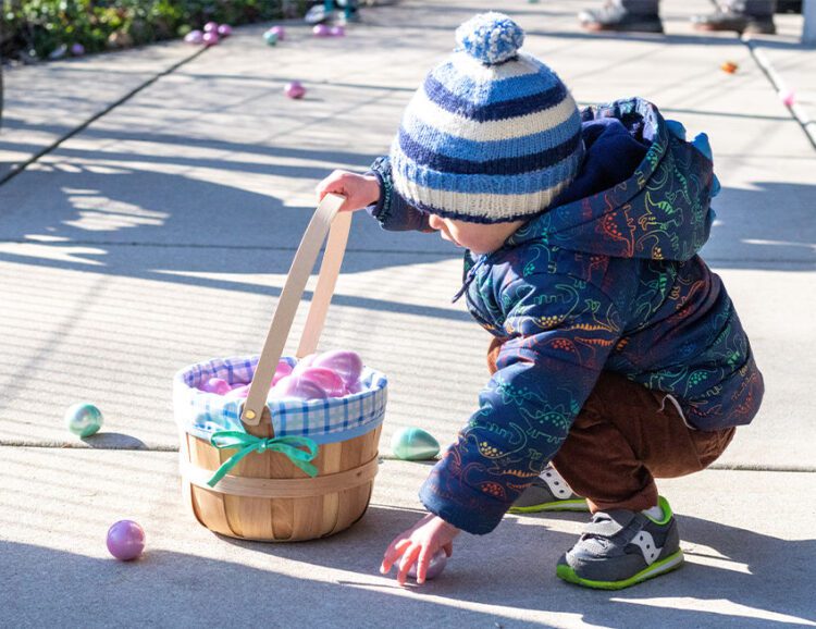 Toddler picking up a plastic egg of the ground with a full Easter basket.